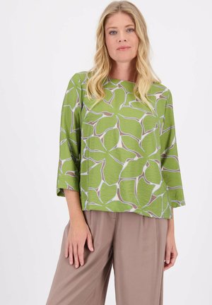 Groene blouse met een bladerenpatroon, met driekwart mouwen en een ronde halslijn, gecombineerd met beige wijde broekspijpen.