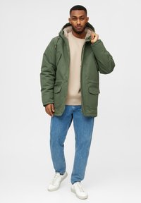 Hombre con cabello corto que lleva una chaqueta de invierno verde con capucha, sudadera beige, jeans azules y zapatillas blancas, de pie frente a un fondo blanco.