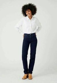 Tamsiai mėlynų aukštai iškeltų džinsų derinys su balta marškinėlių chemise, užbaigtas šviesiai rudos spalvos aulinukais. Audinys su tekstūra ir klasikinis dizainas.