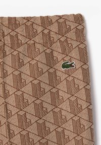 Beige fabric with a repeating geometric pattern in brown and a small green crocodile logo embroidered on the right side.