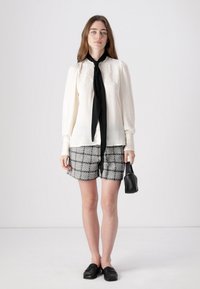 Hvit bluse med blonder og svart slips, kombinert med teksturerte svart-hvite rutete shorts. Svart loafers og håndveske.