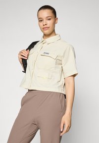 Camicia corta a maniche corte di colore beige chiaro con due tasche frontali, cuciture abbinate e un logo a patch di colore più scuro. Indossata con pantaloni marroni.