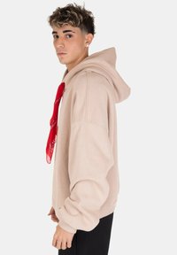 Felpa beige con vestibilità comoda, grande cappuccio e dettaglio in stile bandana con sciarpa rossa. Tessuto morbido e polsini a coste completano il design.