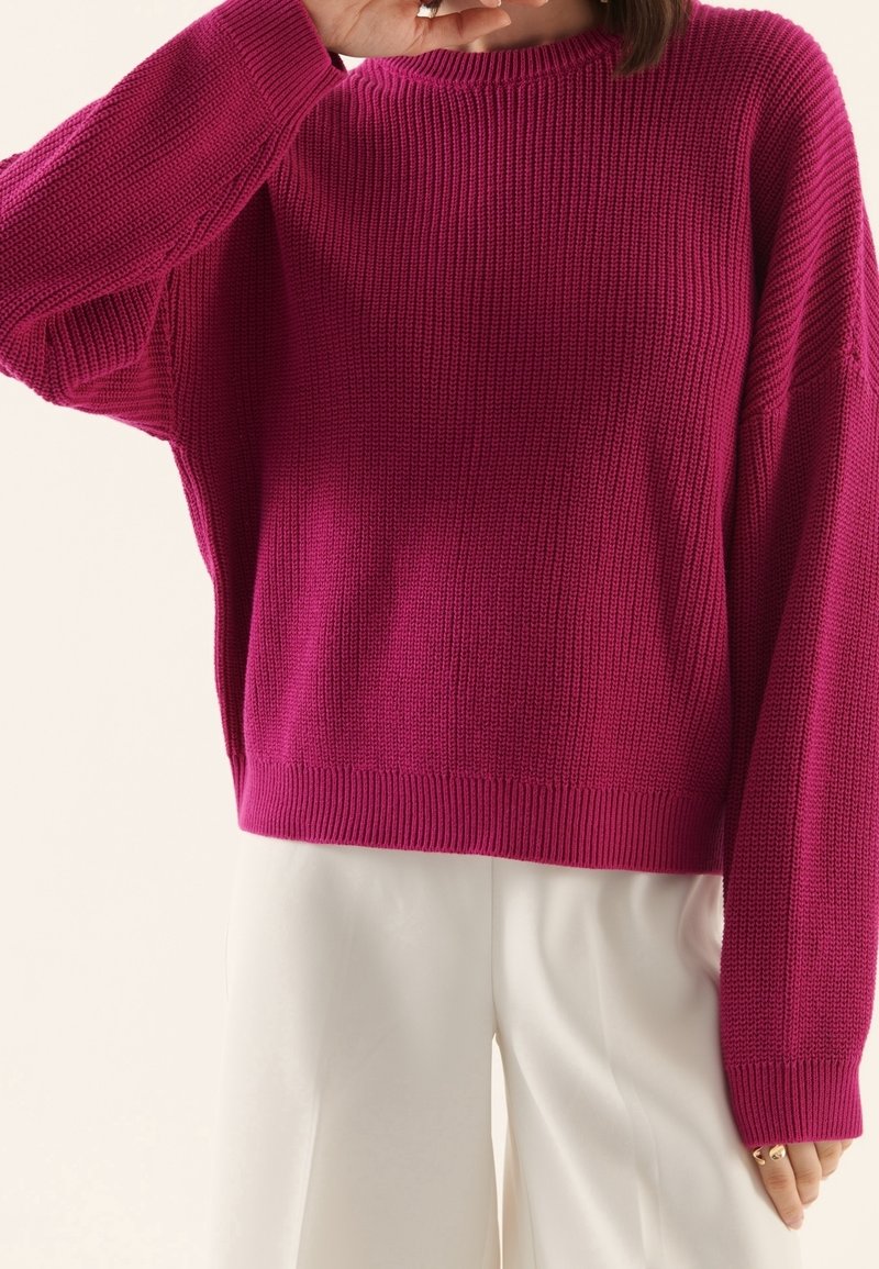 Personne portant un pull en tricot rose vif et un pantalon ample beige clair, une main levée près du visage et l'autre relâchée vers le bas.