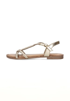 Sandal de oro trenzado con suela plana, que cuenta con una delgada correa para el tobillo y un diseño de punta abierta, exhibida sobre un fondo blanco.