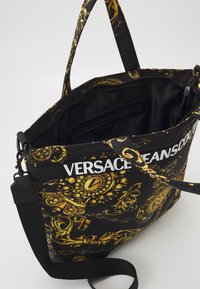 Sac fourre-tout noir Versace Jeans Couture avec motif baroque doré, ouverture supérieure dévoilant une poche intérieure zippée, et bandoulière ajustable.