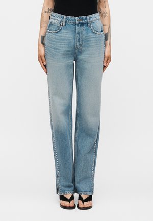 Γυναίκα φορεί ψηλόμεσα ανοιχτό μπλε τζιν με ασημένιες τρουκ κατά μήκος των τσεπών και των πλαϊνών ραφών, συνδυασμένο με μαύρα σανδάλια ανοιχτού δαχτύλου.