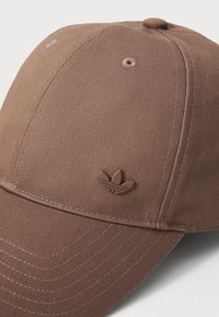 Brun basker med texturerat tyg, med en sydd skärm och en liten präglad logotyp på framsidan. Platt sömnadsdetalj synlig.