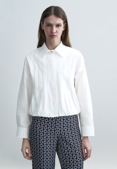Jeune femme portant un chemisier blanc à manches longues et un pantalon bleu marine avec des motifs géométriques blancs et orange, debout devant un fond gris clair.