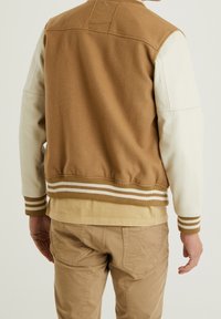 Brun ullblandad bomberjacka med vita läderslag, randiga ribbade manschetter och en lätt lyster. Neutral beige skjorta och kaki byxor synliga.