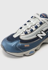 Blå och silver sportskor med nätpaneler, gummisula och "1000"-logotyp. Har en vadderad plös och snören med reflexdetaljer.