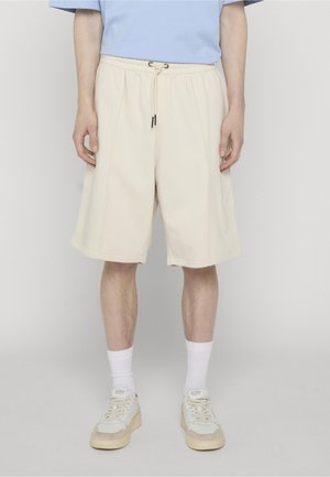 Person som har på seg beige knelengde shorts med snor, hvite crew-sokker og kremfargede joggesko, iført en lyseblå skjorte som er kuttet ut over midjen.