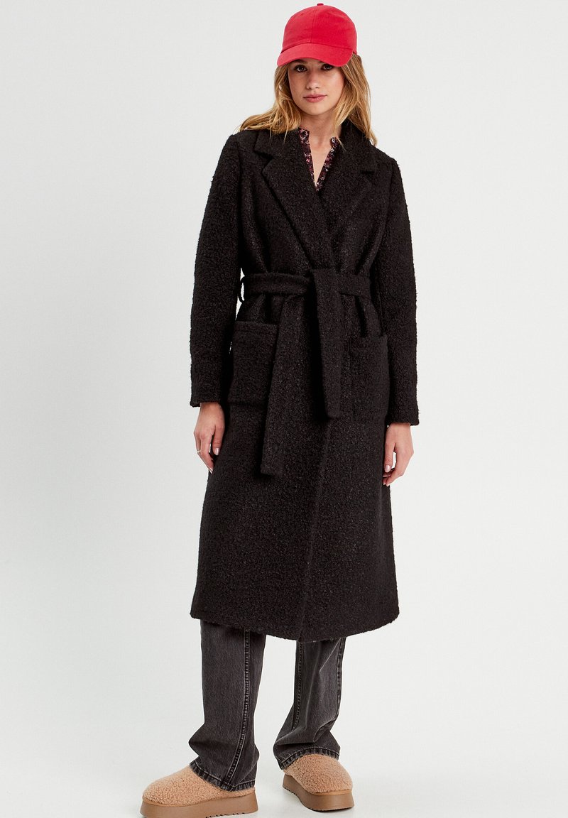 Black long coat made of textured material, featuring a belt, two front pockets, and a notched collar, worn over dark jeans and beige slippers.