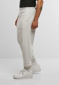 Muž v svetlosivých teplákach so bočnými vreckami, bielych ponožkách a bielych teniskách Nike stojí na jednoliatom pozadí.