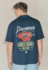 Mørkeblå oversize T-shirt med kortærmer, der har et grafisk print af tomater og tekst, med en afslappet pasform og blød stofstruktur.