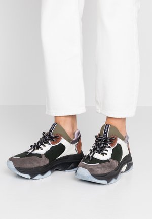 Grube sneakersy w kolorach czarnym, białym, szarym, brązowym i oliwkowozielonym z wzorzystymi sznurowadłami, noszone z białymi, krótkimi spodniami na szarej podłodze.