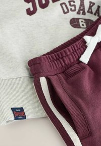 Pantaloni della tuta bordeaux con strisce laterali bianche e vita elastica, abbinati a una felpa grigia chiara con testo bordeaux e etichetta navy Superdry.
