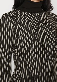 Femme portant une robe à manches longues à motifs noir et blanc avec un col haut noir, vue du cou à la taille.