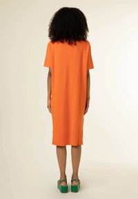 Personne aux cheveux bouclés portant une robe orange jusqu'aux genoux et des sandales compensées vertes, debout de dos sur un fond uni.