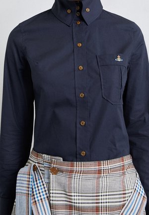 Mørk marineblå skjorte med brune knapper og pocketlogo, stoppet ned i flerfarvet ternet nederdel med overlappende paneler og knapdetaljer.