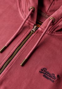 Sudadera con cremallera color marrón burdeos y acabado texturizado, con herrajes en tono dorado, cordones ajustables y logotipo de la marca bordado cerca de la cremallera.