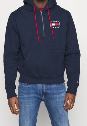 Mężczyzna noszący granatową bluzę z kapturem Tommy Jeans z czerwonymi troczkami i logo na piersi, w połączeniu z niebieskimi jeansami na jednolitym tle.