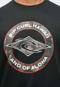 Sort T-shirt i bomuld med et cirkulært grafisk motiv med teksten "RIP CURL HAWAI'I" og "LAND OF ALOHA," fremhævet med et camouflage-mønster.