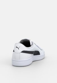 Baskets basses blanches avec une bande logo Puma noire et un emblème chat Puma noir sur le talon, inclinées pour montrer l'arrière et le côté.