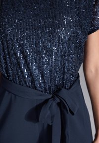 Navy sequins top with a textured surface, short sleeves and a tied waist sash in matching fabric. Smooth and sleek finish is visible.