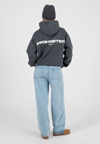 Hallhalli kapuutsiga dressipluus, mille seljal on toonikalt valgete suurte tähtedega kiri "KEELATUD", kantud helesiniste lahtiste teksade ja pruunide saapadega.