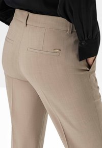 Beige maßgeschneiderte Hosen mit glatter Textur, ausgestattet mit zwei Gesäßtaschen und goldfarbenen Hardware-Akzenten. Kombiniert mit einer schwarzen Bluse.