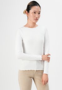 Young woman with hair in a bun wearing a fitted white long-sleeve top and beige corduroy pants, holding one arm with the opposite hand.