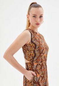 Vestido sin mangas con una tela marrón con patrones geométricos en naranja, crema y azul, con cortes laterales y bolsillos.