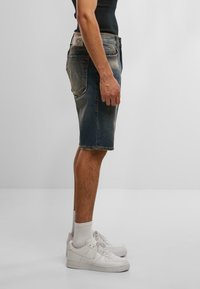 Muž stojaci v vyblednutých modrých džínsoch, bielych ponožkách, bielych teniskách a čiernej bezrukávovej košeli, zobrazený z bočného pohľadu na jednofarebnom pozadí.