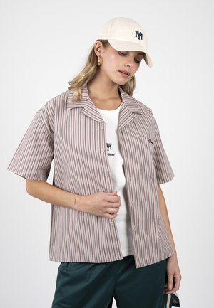 Nő fehér sapkában, függőleges csíkos rövid ujjú gombos ing fehér top fölött, sötétzöld rövidnadrágban, lefelé néz.