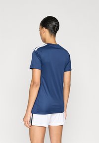 Αθλητική μπλούζα σε ναυτικό μπλε με κοντά μανίκια, με λευκές ρίγες στους ώμους, κατασκευασμένη από απαλό ύφασμα που απομακρύνει την υγρασία. Συνδυάζεται με λευκό σορτς.