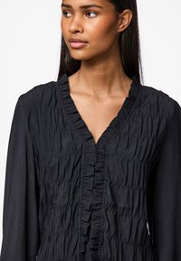 Čierne šatové šaty s nariasenou textúrou, výstrihom do V a volánovým predným dielom. Dlhé rukávy a plynulý strih umocňujú ich siluetu.