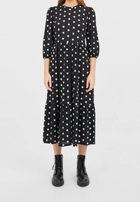 Zwarte midi-jurk met witte polka dots, ronde halslijn, driekwart mouwen, geplooide zoom, gecombineerd met zwarte enkellaarzen.