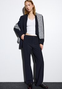 Jonge vrouw in een marineblauw gestreept pak met brede broekspijpen en blazer over een witte V-hals top, grijze sjaal over de schouders gedrapeerd, zwarte instappers.