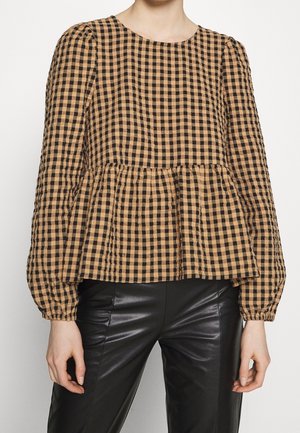 Kobieta w brązowo-czarnej, kraciastej bluzce z długim rękawem, marszczonym mankietem i dołem w stylu peplum, w połączeniu z czarnymi skórzanymi spodniami.