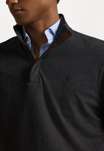 Μαύρο πουλόβερ σε σχέδιο ρόμβου με φερμουάρ έως το ένα τέταρτο, καφέ σουέτ γιακά και ανοιχτό μπλε πουκάμισο από κάτω, με υφή ύφασματος και διακριτική λεπτομέρεια λογότυπου.