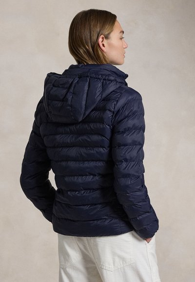 Pruunide juustega naine, kes kannab tumesinist tikandiga kapuutsiga jope ja valgeid pükse, seisab käed taskutes, näoga paremale.