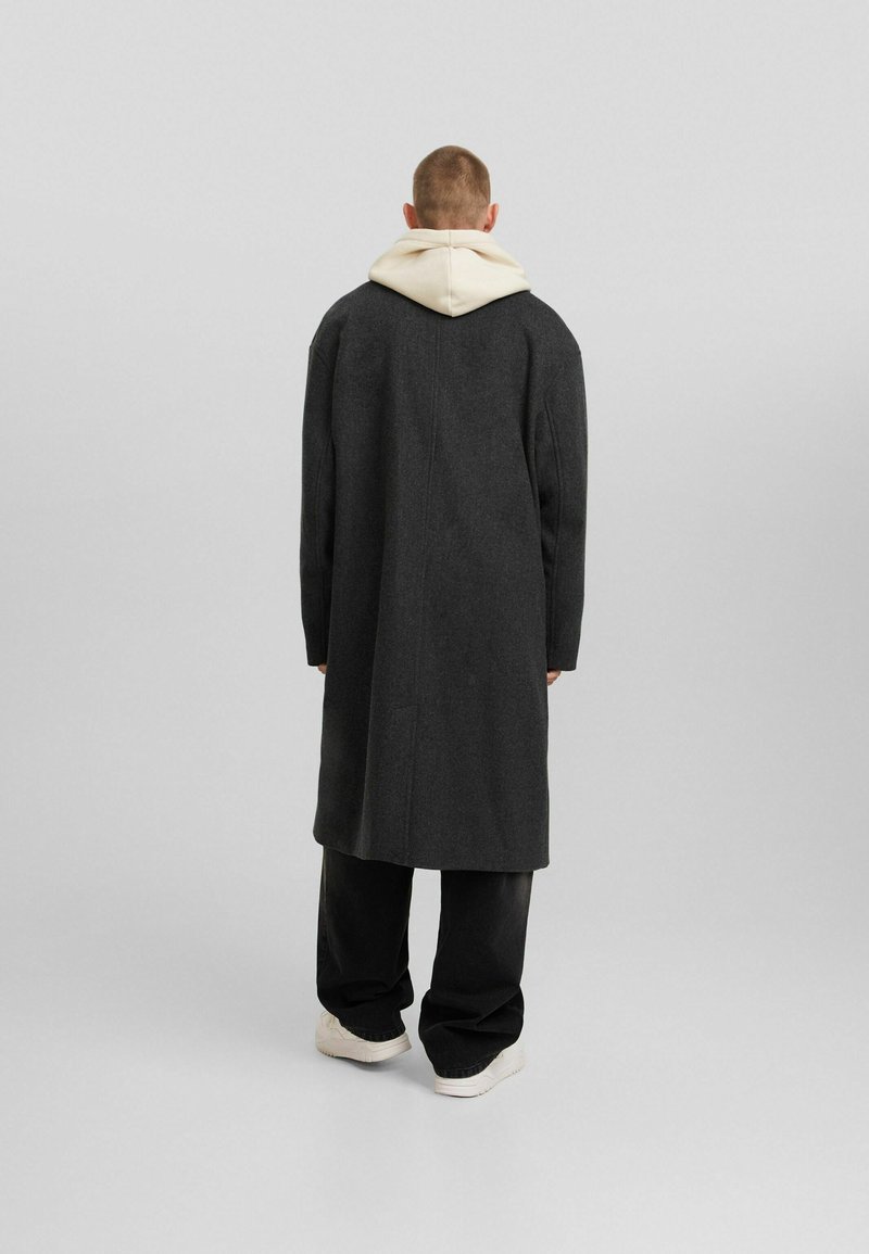 Lang, donkergrijze jas van een wolblend, met een capuchon en een rechte snit, gecombineerd met losse zwarte broeken en sneakers.