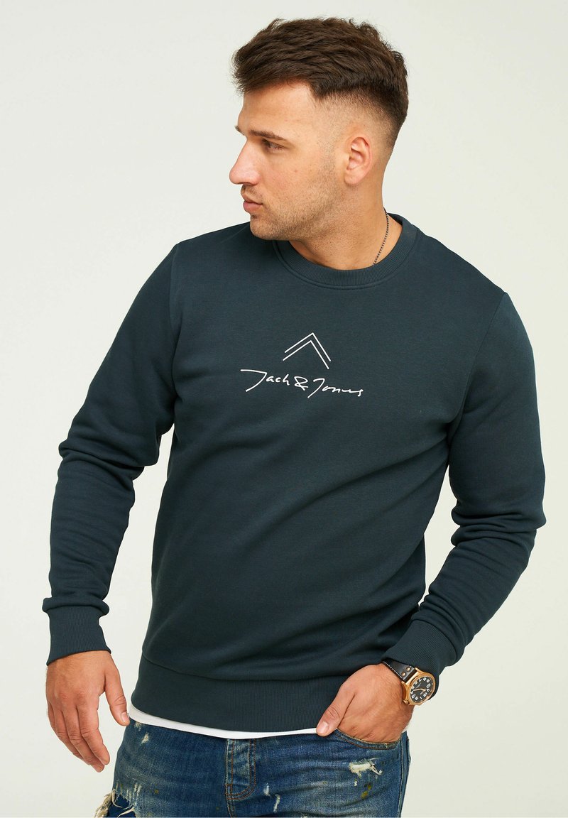 Dark green sweatshirt with long sleeves, ribbed neckline and cuffs, featuring a white logo print on the chest. Worn over distressed blue jeans.