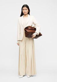 Γυναίκα με κρεμ σακάκι και παντελόνι φαρδιάς γραμμής, κρατώντας μια μεγάλη καφέ δερμάτινη τσάντα φάκελο, στέκεται σε απλό λευκό φόντο.