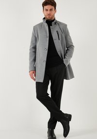 Bărbat îmbrăcat cu un palton gri, cu nasturi, peste un pulover negru cu guler înalt, pantaloni negri și cizme Chelsea negre, stând cu o picior încrucișat.