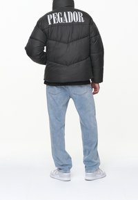 Svart puffjacka med hög krage och "PEGADOR" i stora grå bokstäver på ryggen, parat med ljusblå jeans och grå sneakers.