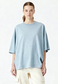 Světle modré oversized tričko vyrobené z měkké texturované látky. Má kulatý výstřih a volné rukávy, v kombinaci s béžovými cargo kalhotami.