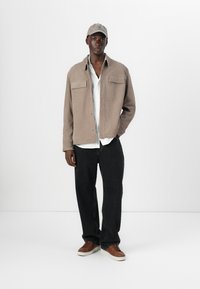 Beige suede jacket with two chest pockets, white shirt beneath, black wide-leg jeans, and brown shoes. Simple cap completes the outfit.