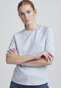 Bluză ușor albastră cu mânecă scurtă, cu decolteu rotund și mâneci raglan, realizată dintr-un material neted cu o textură subtilă și cusături curate.